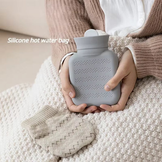 Person holding a gray silicone hot water bag with a matching textured cover on a soft surface.